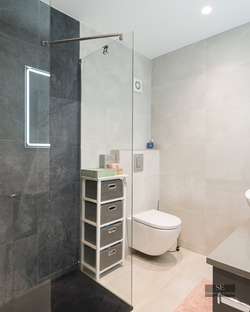 Salle de bain moderne avec douche en verre, carrelage gris foncé, vasque blanche et toilettes suspendues.
