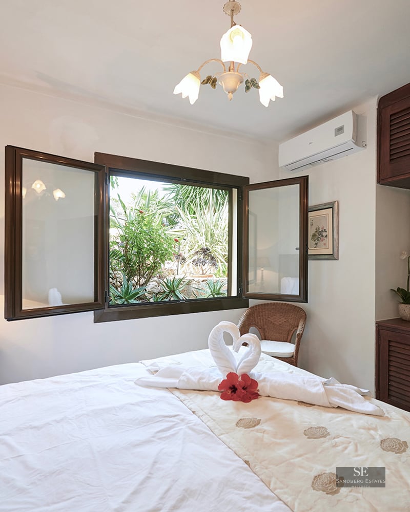 Bright bedroom with white linens, dark wood closets, and an open window looking onto a lush green garden.