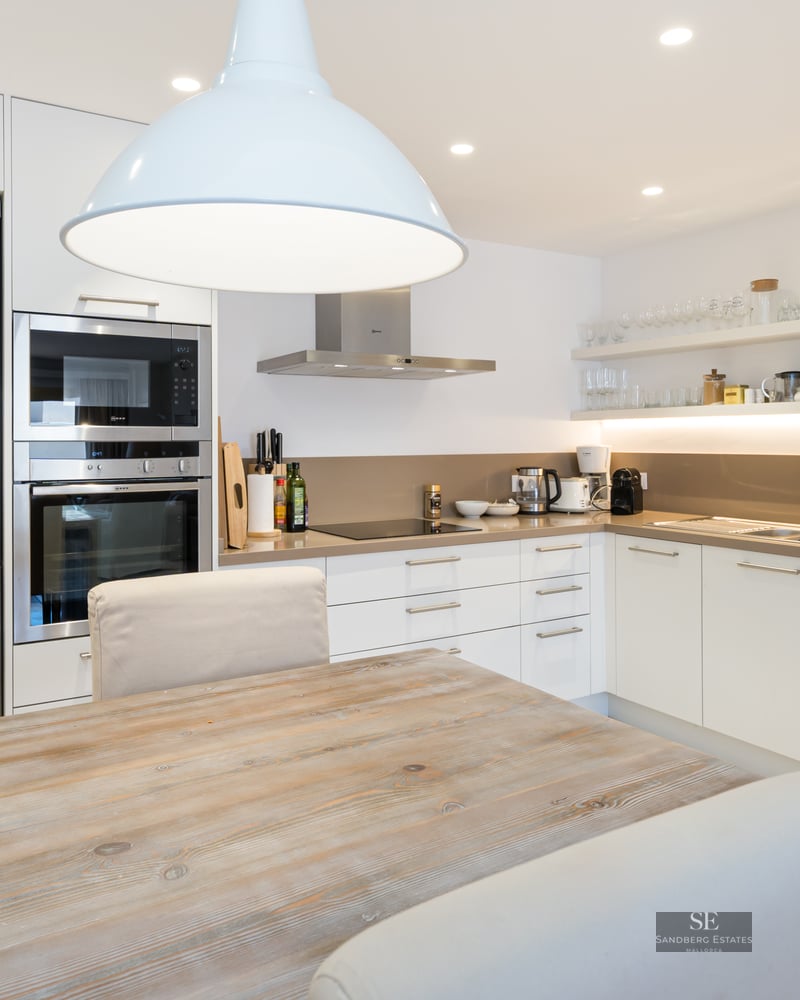 Modern white kitchen with stainless steel appliances, a wooden table, and beige upholstered chairs under a pendant light.