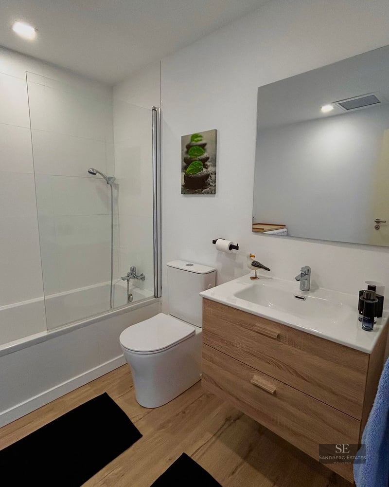 Contemporary bathroom with white walls, glass-screened bathtub, toilet, and light wood vanity unit.