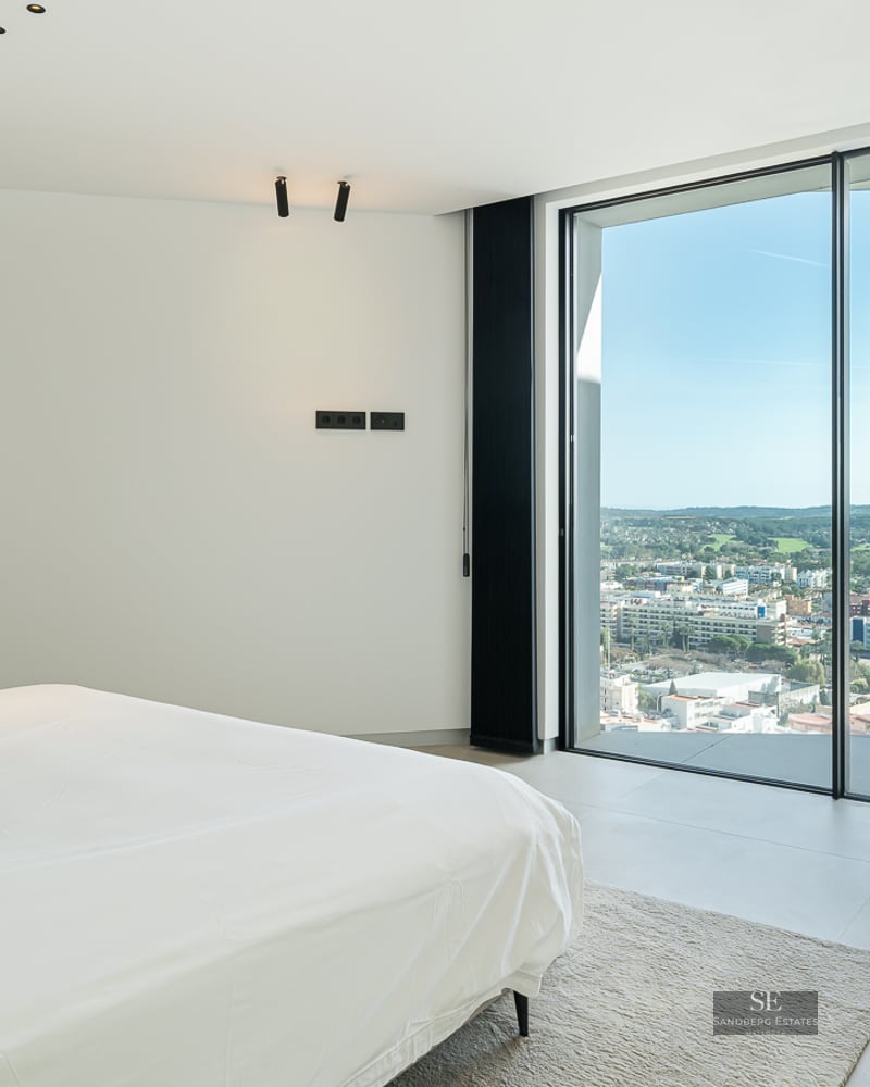 Minimalist bedroom featuring a white bed, grey armchair, and floor-to-ceiling windows overlooking a city cityscape.