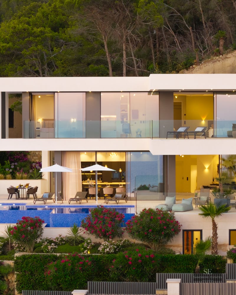 Facade of a modern white two-story villa with an infinity pool and large glass windows at dusk.