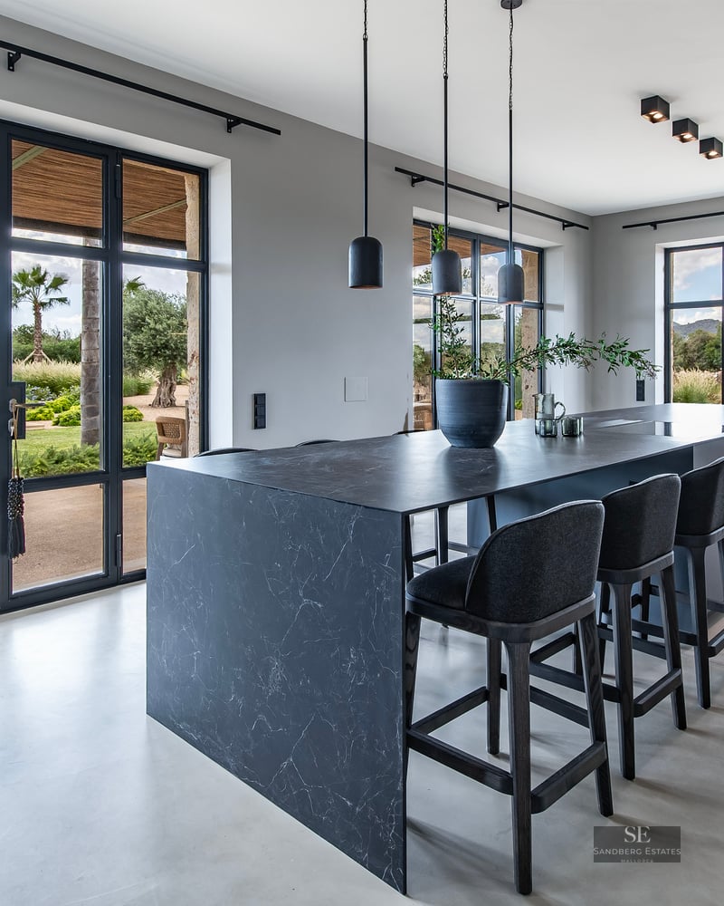 Modern kitchen featuring a large dark marble island, black stools, and glass doors opening to a garden and pool.