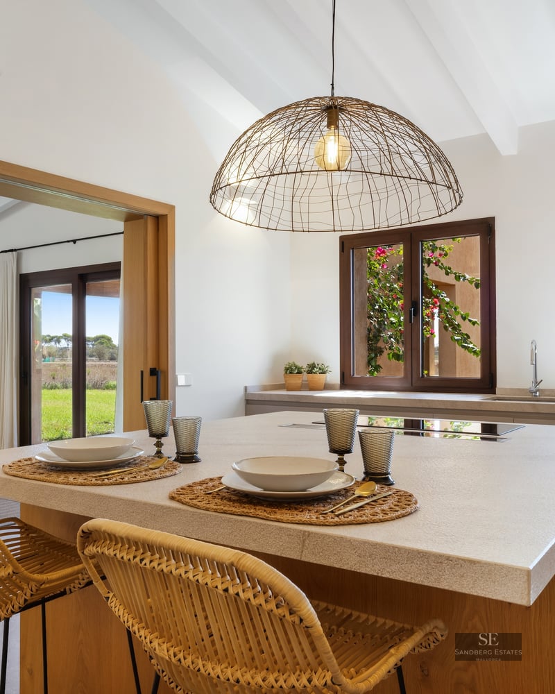 Modern kitchen island with wicker stools and white beamed ceiling overlooking rural landscape.