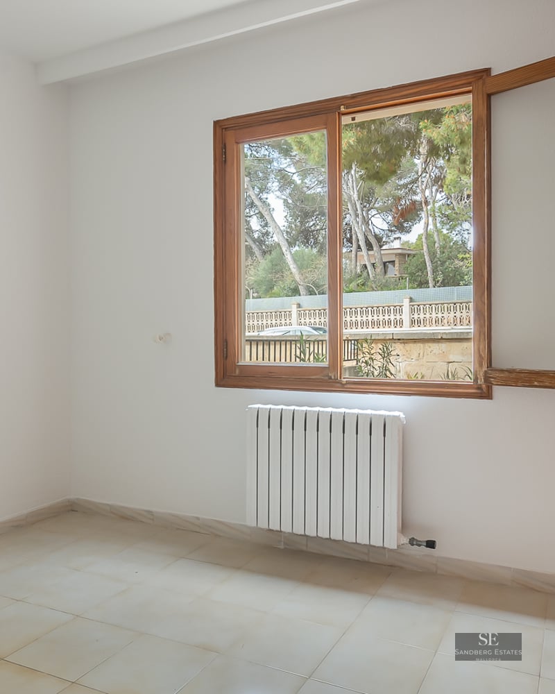 Empty bedroom with white walls, tiled floor, wooden built-in wardrobe, and window overlooking trees.