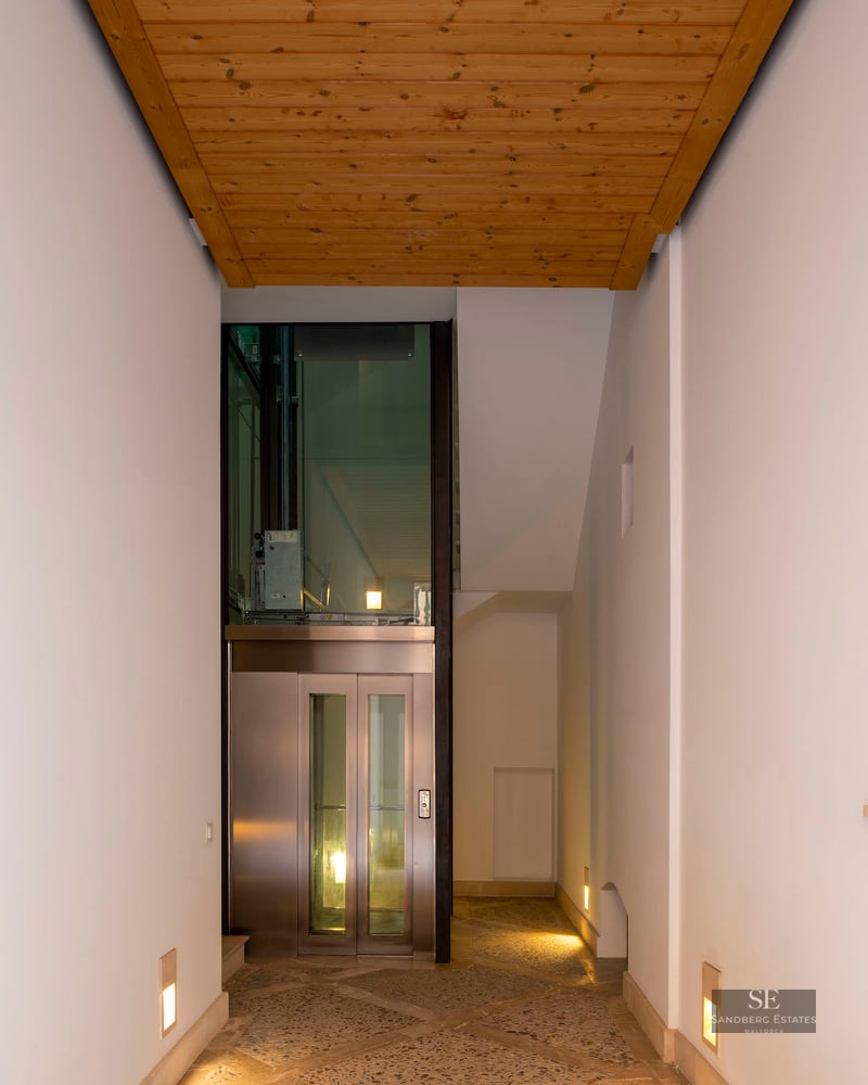 Modern hallway with stone mosaic flooring, wood ceiling, and a glass elevator at the end.