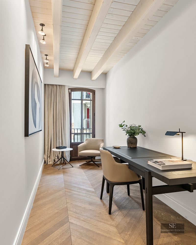 Modern home office with dark desk, chevron wood floor, white beamed ceiling, and a large window.