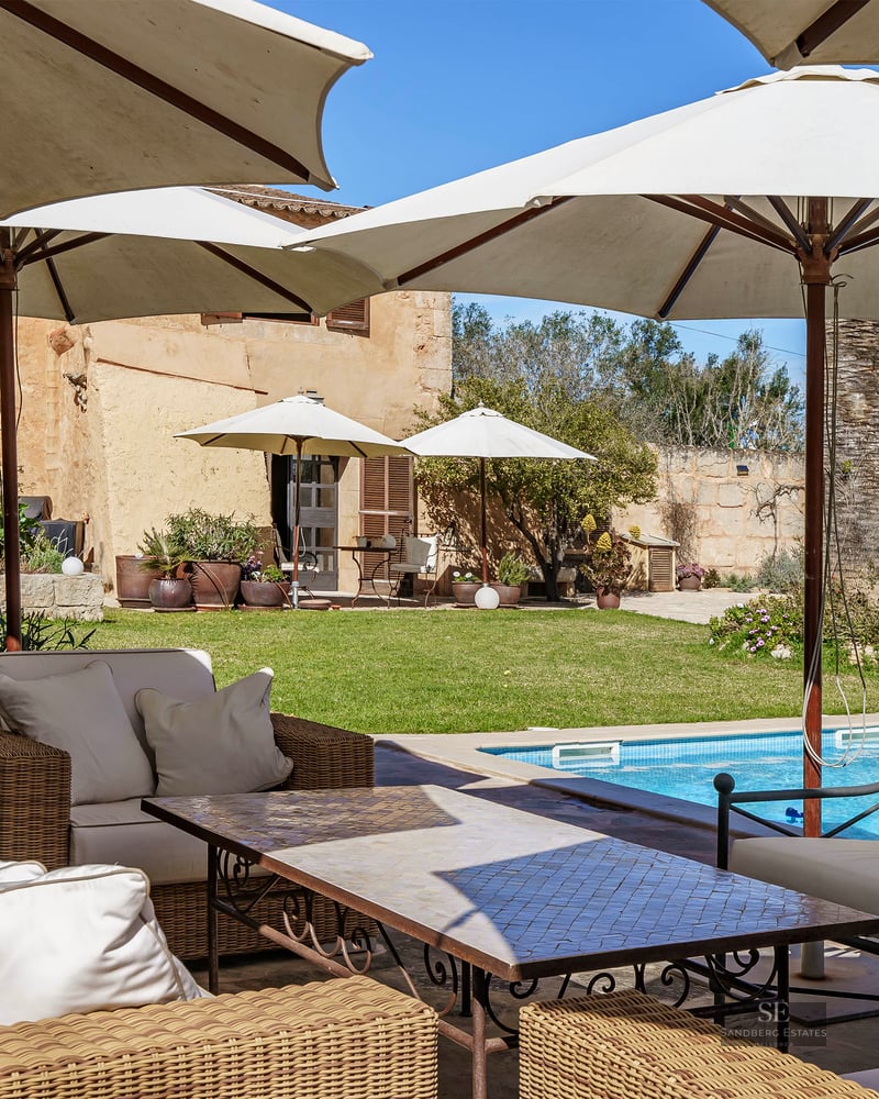 A luxury swimming pool surrounded by large white umbrellas and wicker lounge furniture in a Mediterranean garden.