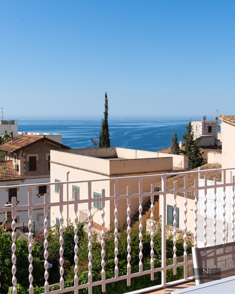 Panoramic view of the blue sea over coastal rooftops from a white balcony with outdoor seating.