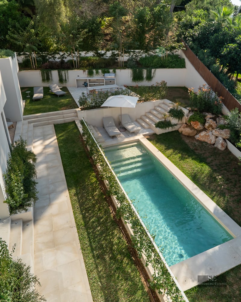 High-angle view of a rectangular turquoise pool surrounded by lush gardens and a modern white villa with green roofs.
