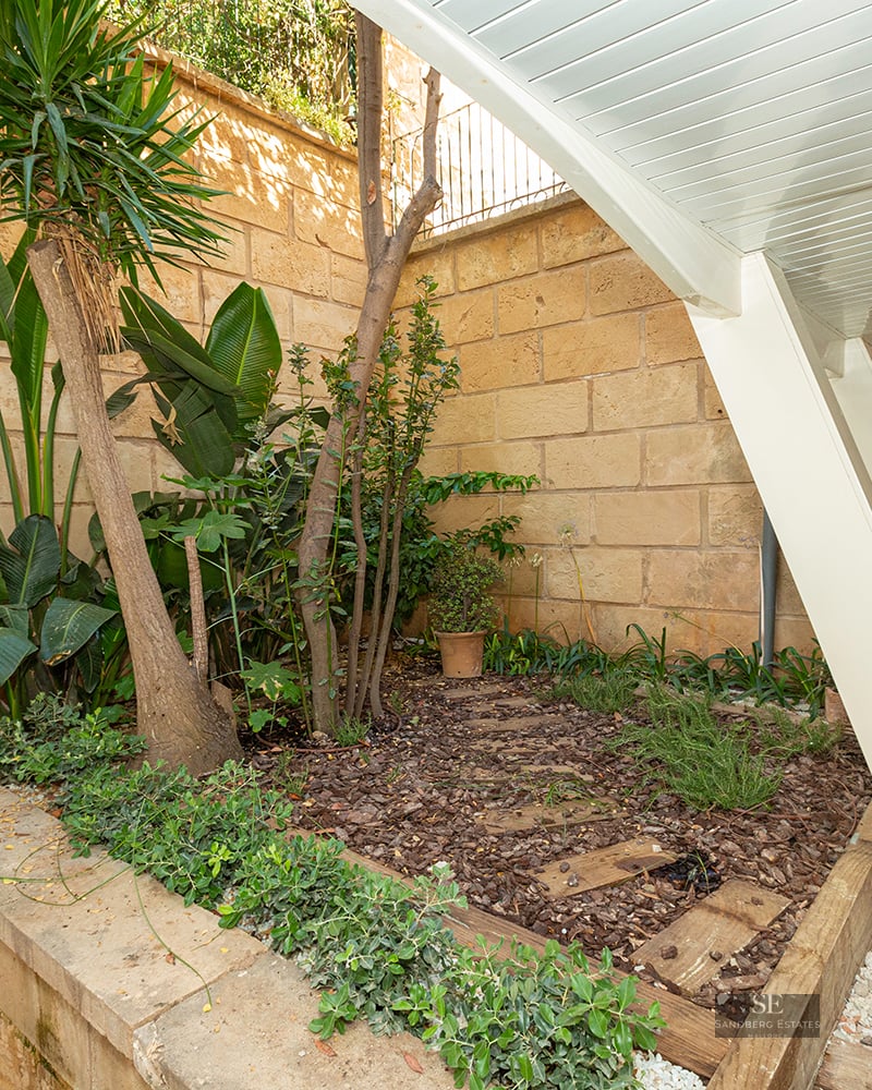 A small outdoor garden area with tropical plants, wood mulch, and a sandstone wall under a slanted white structure.