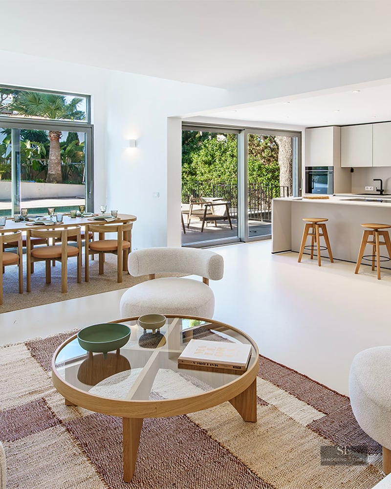 Bright open-plan room with modern white sofas, wood dining set, and minimalist kitchen overlooking a pool and garden.
