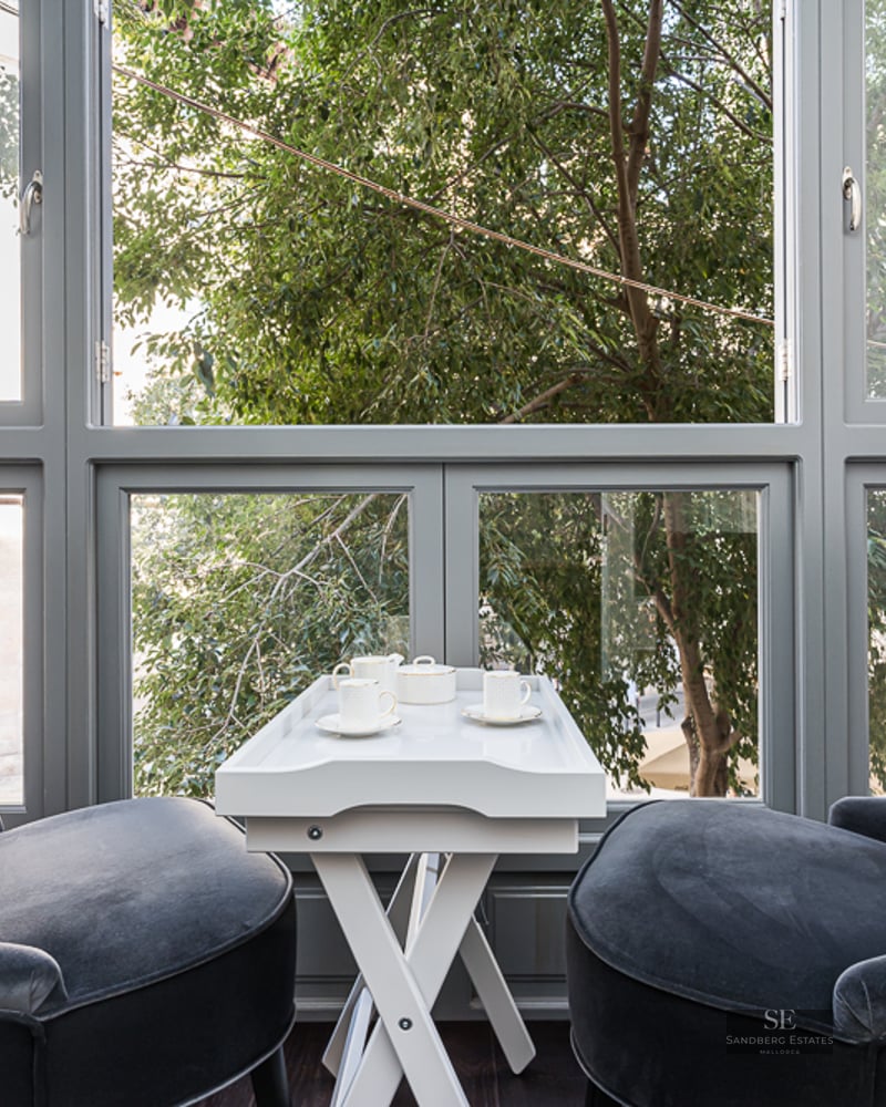 Two charcoal velvet armchairs facing a large window with a white tea table in between, overlooking city trees.