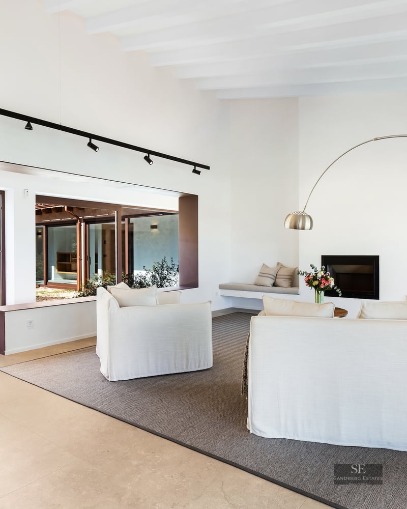 Bright living room with white sofas, an arched floor lamp, a fireplace, and views of an olive tree in a courtyard.