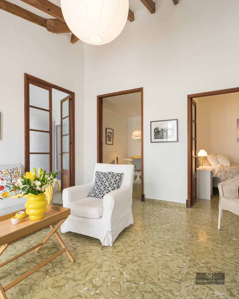 Bright living room with white sofa, rustic wooden ceiling beams, patterned green floor tiles, and doorways to other rooms.