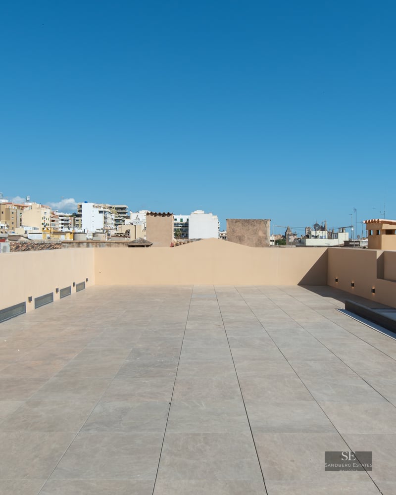 Spacious rooftop terrace with beige tiles and modern wall lighting under a clear blue sky.