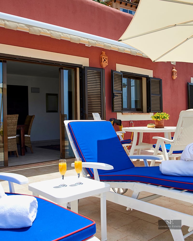 Private terrace with blue sun loungers, white towels, orange juice, and an umbrella against a terracotta building.