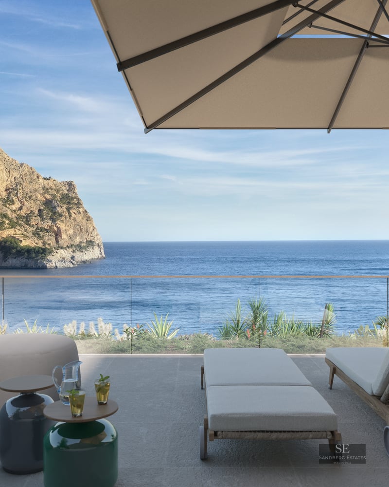 A sunny terrace with sun loungers, a parasol, and a glass railing overlooking the Mediterranean sea and a rocky cliff.