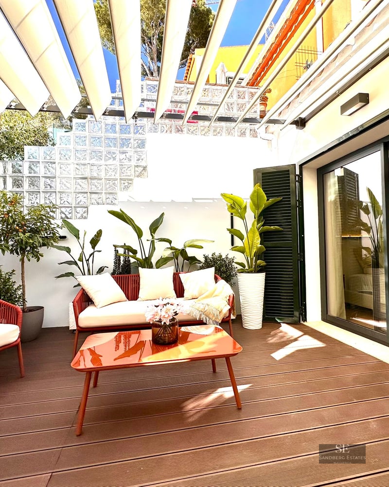 Stylish outdoor terrace with red and white furniture, orange table, lush plants, and a white retractable awning in the sun.