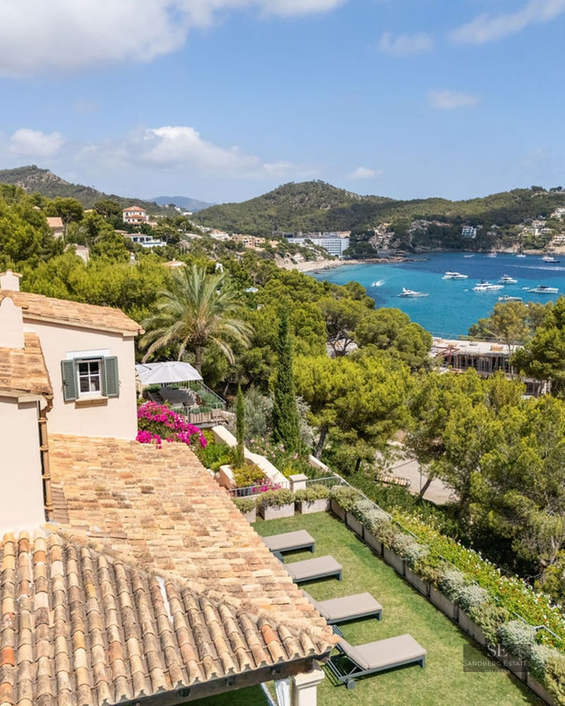 High-angle view of a villa with a tiled roof overlooking a garden, a turquoise bay with boats, and lush hills.
