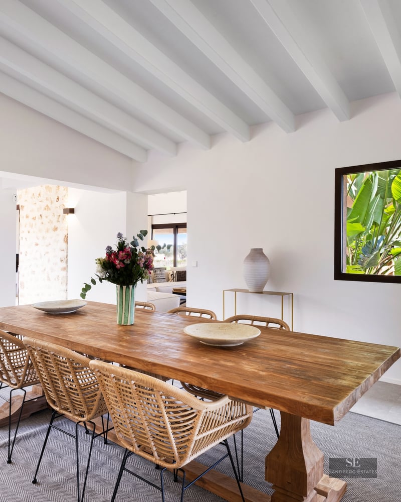 Bright dining room with a large wooden table, wicker chairs, white beamed ceiling, and lush garden views.
