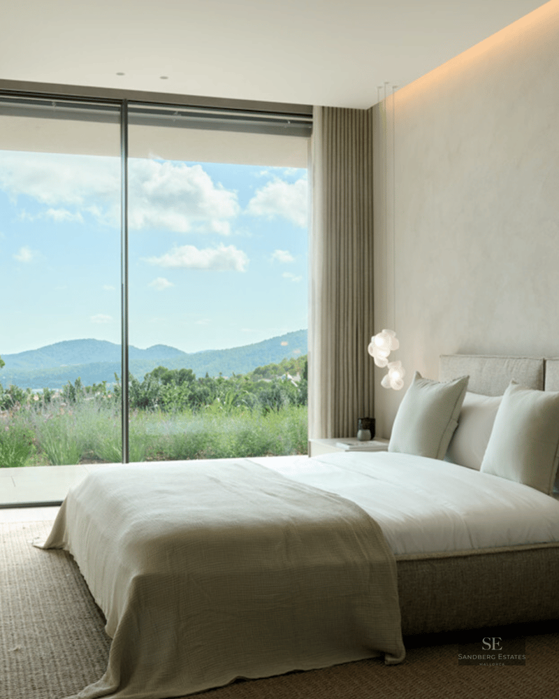 Luxury bedroom featuring textured walls, a minimalist bed, and floor-to-ceiling windows overlooking hills.