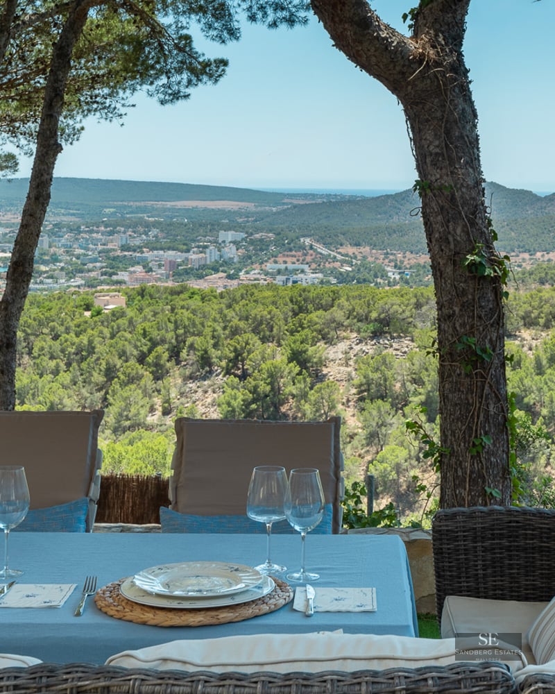 Outdoor dining table and wicker sofa on a shaded terrace overlooking a coastal town and sea.
