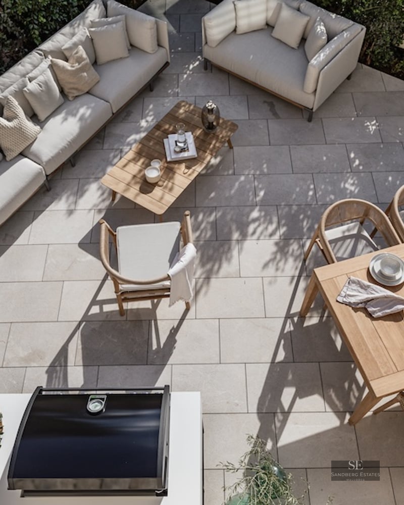 High-angle view of a stone-paved terrace with designer sofas, wooden dining table, and outdoor kitchen area.