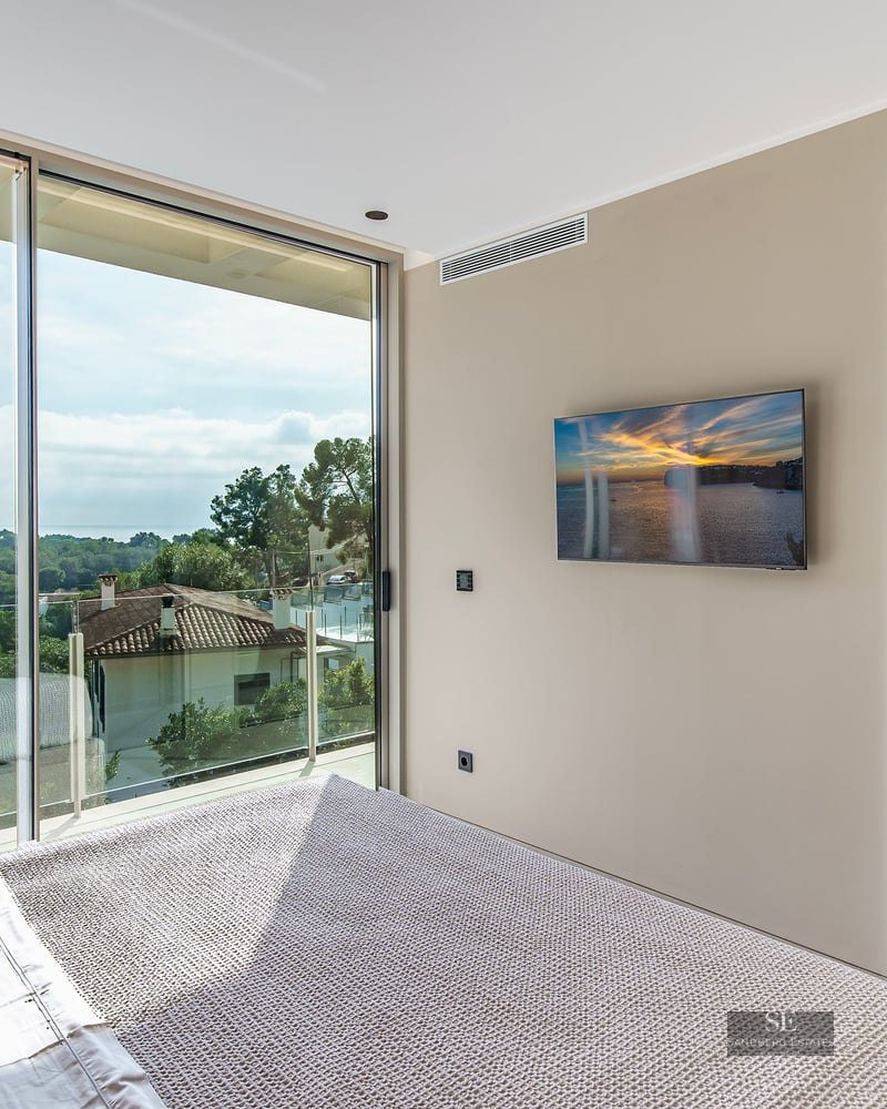 A modern bedroom with a large floor-to-ceiling window overlooking a forest and sea, with an ensuite bathroom visible.