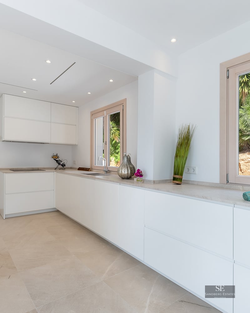 Bright, modern kitchen featuring white handleless cabinets, stone countertops, and windows overlooking greenery.