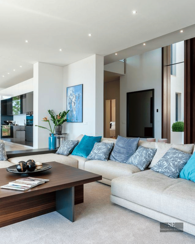 Spacious white living room with a large gray sectional sofa, teal pillows, dark wood accents, and an open kitchen view.