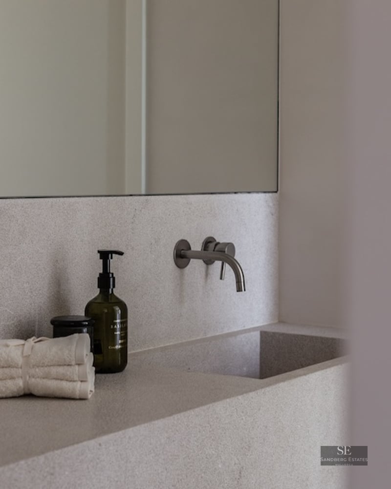 Close-up of an integrated light stone bathroom sink with a wall-mounted brushed metal faucet and luxury amenities.