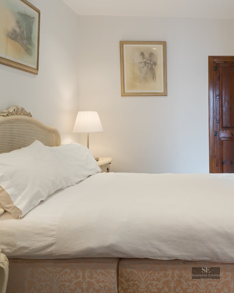 A cozy bedroom featuring a white bed with a cane headboard, twin bedside lamps, and a dark vintage wooden door.