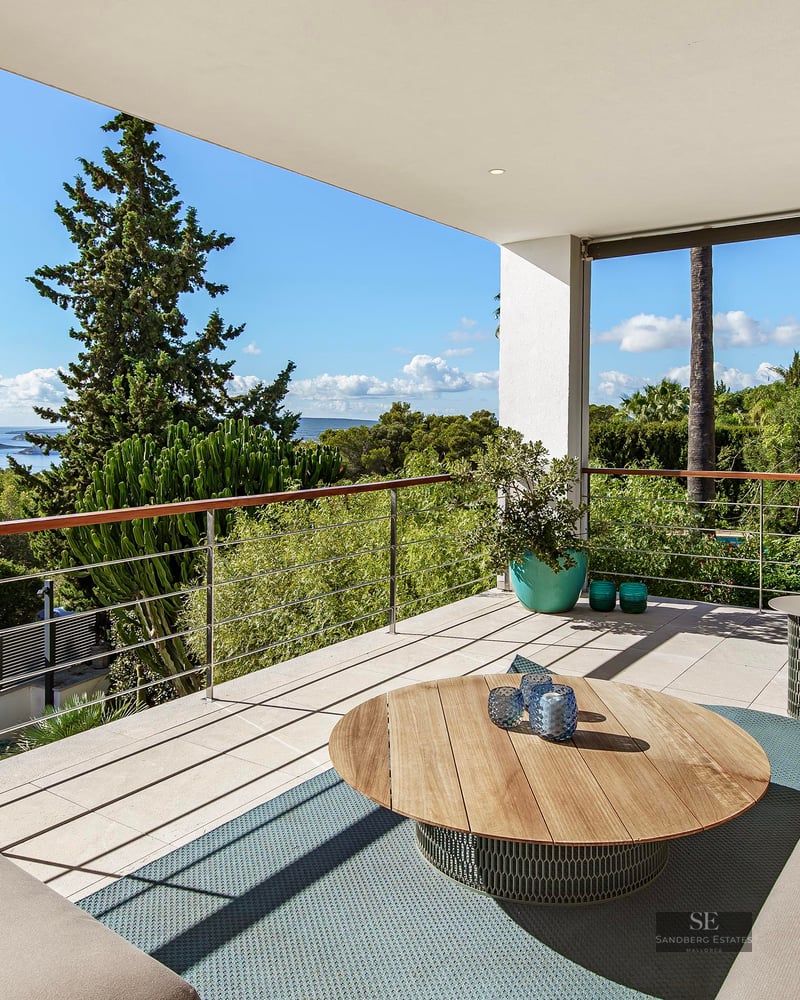 Terrasse moderne avec canapés beiges, table en bois et garde-corps métallique surplombant la mer et la verdure.