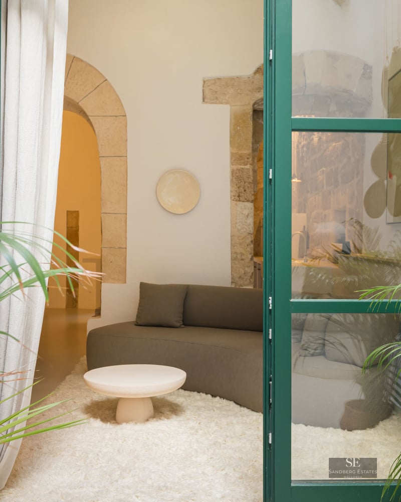 View through green glass doors into a living room with a grey sofa, white shaggy rug, and stone archway.