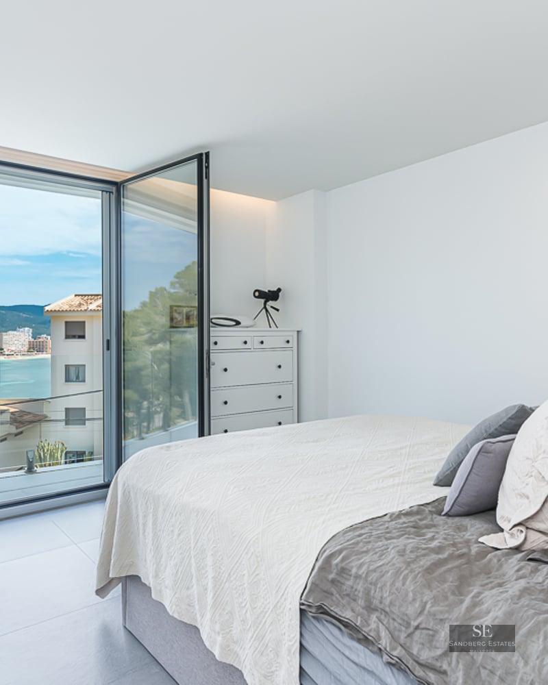 Bright bedroom with a bed and large glass doors opening to a coastal bay view and mountains.
