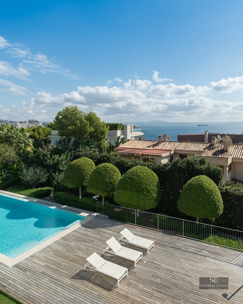 Elevated view of a rectangular turquoise swimming pool next to a wooden deck with white sun loungers and ocean views.