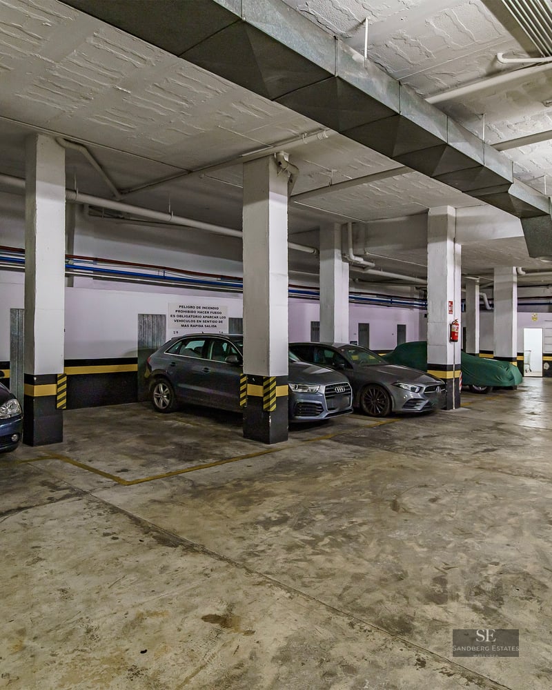 Grand garage souterrain avec piliers en béton, marquages de sécurité jaunes et voitures garées.