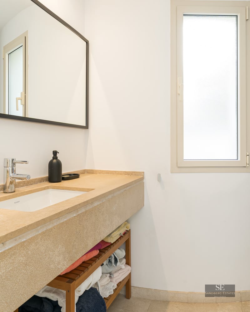 Modern bathroom featuring a long beige stone vanity, large black-framed mirror, and glass shower door in a bright room.