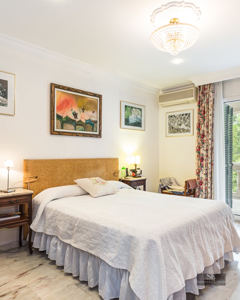 Elegant bedroom with a double bed, marble flooring, crystal chandelier, and glass doors opening to a balcony.
