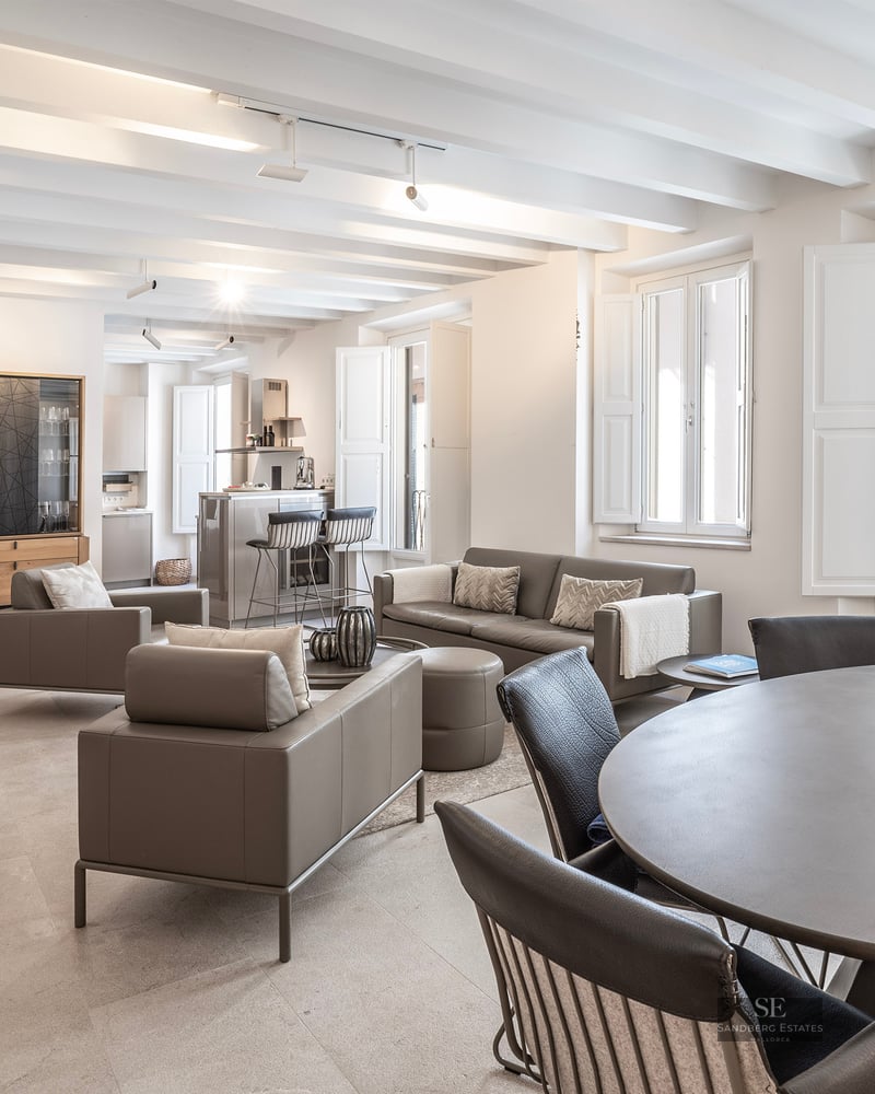 Elegant open-plan living room with gray sofas, white beamed ceiling, and natural light from large shuttered windows.