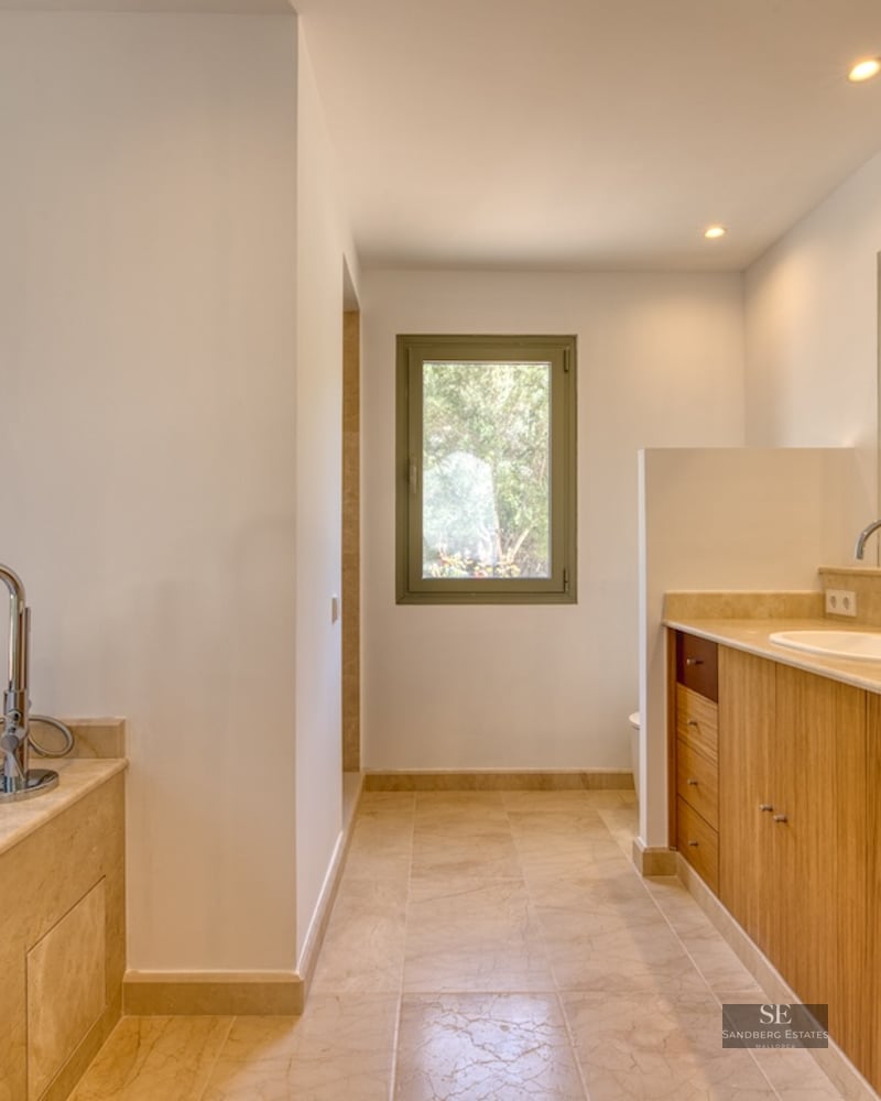 Luxury bathroom featuring wooden double vanity, beige marble countertops, bathtub, and natural light.