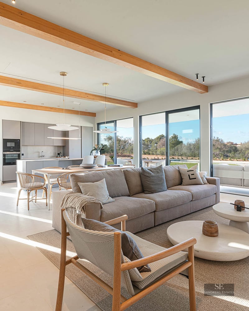 Spacious modern living room with large beige sofa, wooden ceiling beams, and floor-to-ceiling glass doors.