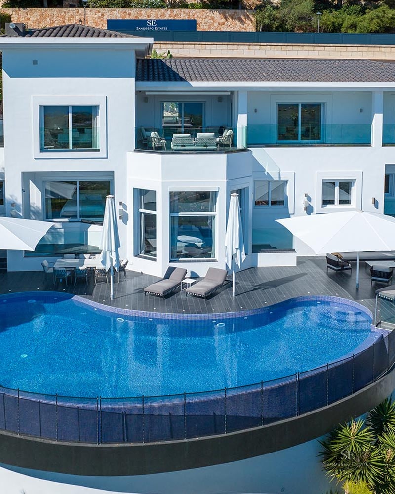 Elevated view of a modern white villa featuring a large blue semicircular infinity pool and furnished terraces.
