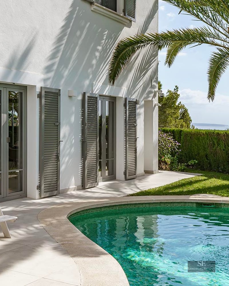 A turquoise swimming pool beside a white villa with a palm tree, grey shutters, and a distant ocean view.