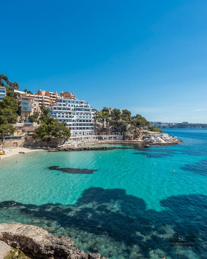 Wide panoramic view of a crystal-clear turquoise bay with a sandy beach and white buildings on a rocky coastline.