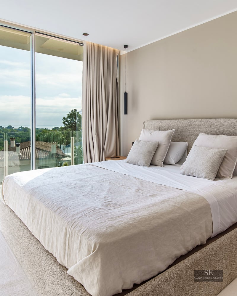 Modern master bedroom featuring floor-to-ceiling glass walls overlooking a lush green landscape with a large neutral bed.