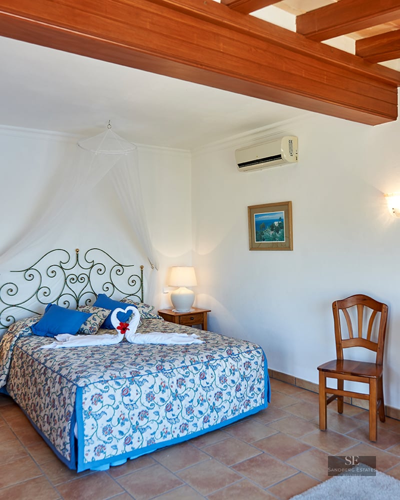 Bedroom with terracotta tile floor, exposed wooden ceiling beams, wrought iron bed, and classic wooden furniture.