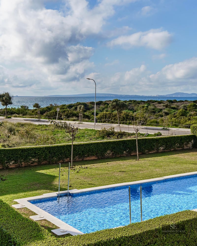 Rectangular blue pool surrounded by green lawn and hedges with the Mediterranean sea in the background under cloudy sky.