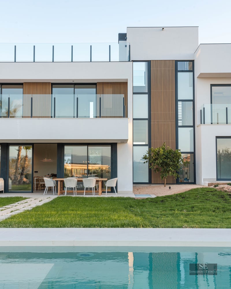 Modern white villa with wood accents, large windows, and a turquoise swimming pool in front of a green lawn.